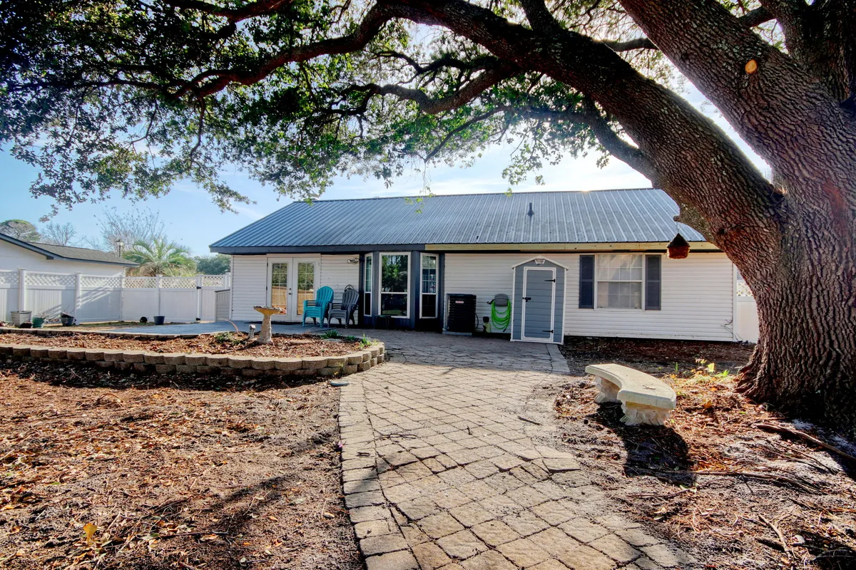A welcoming backyard scene featuring a sprawling oak tree as the central focal point. The stone pathway guides you beneath the tree’s canopy, leading to an adjoining seating area perfect for gatherings. The surrounding landscaping is lush, with mature greenery and glimpses of the house’s exterior, all bathed in soft natural light.