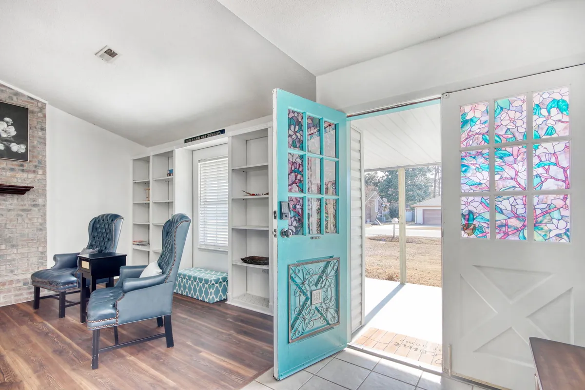 This image features a turquoise door with a stained glass window, flanked by comfortable chairs on wooden flooring. The welcoming mood is enhanced by the natural light and decor, suggesting a peaceful entryway or sunroom where one might pause and relax before stepping outside or returning home.