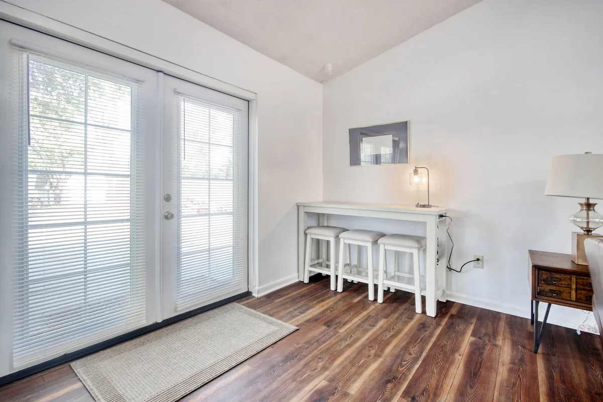 A bright, inviting space with double doors that feature blinds, a bar table with stools set for casual dining, a table lamp, and a mirror reflecting light. The scene suggests a cozy nook for morning coffee or relaxing after a day exploring the coast, with views through the doors hinting at the outdoors.
