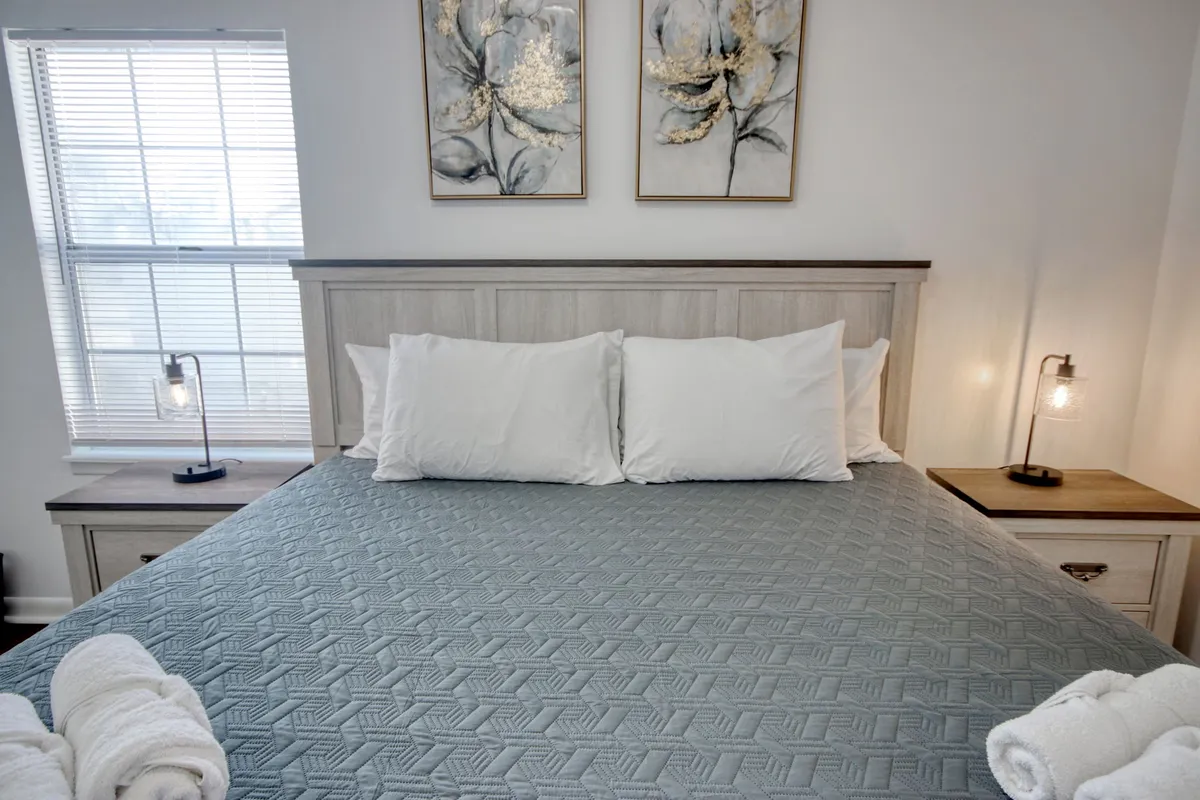 A bright and serene bedroom featuring a neatly made bed adorned with decorative pillows, floral artwork above the headboard, and a nightstand with a lamp. Natural light fills the room, creating a calm atmosphere perfect for rest and relaxation.