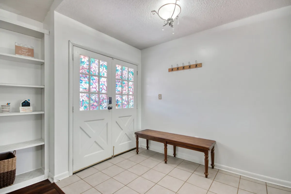 The image shows a sunlit entrance area with a stained glass door, a wooden bench for sitting or removing shoes, and a shelving unit for storage. The inviting mood suggests a space where guests and residents are warmly welcomed, and where one prepares to step out into the neighborhood.
