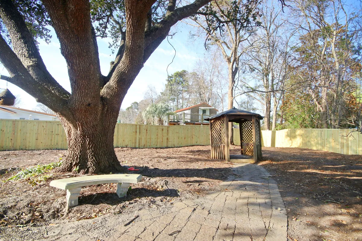 A tranquil garden scene with a large shaded tree at its center, a charming wooden gazebo tucked nearby, a stone pathway, and a wooden bench. The space is alive with possibilities for relaxation, reading, children’s play, or intimate outdoor gatherings.