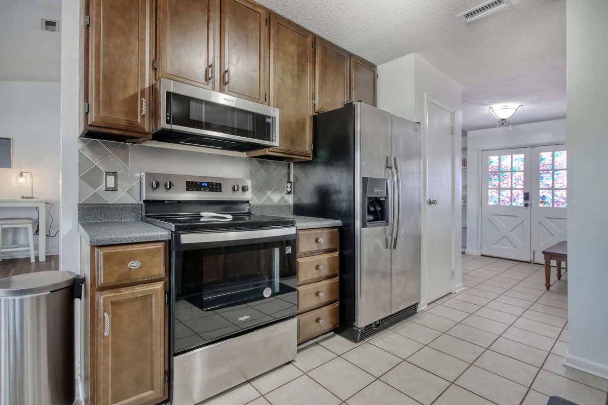 A functional kitchen scene with stainless steel appliances, wooden cabinets, tile flooring, and an adjacent dining area filled with natural light. The setup highlights the kitchen's modern amenities and its suitability for both cooking and socializing.