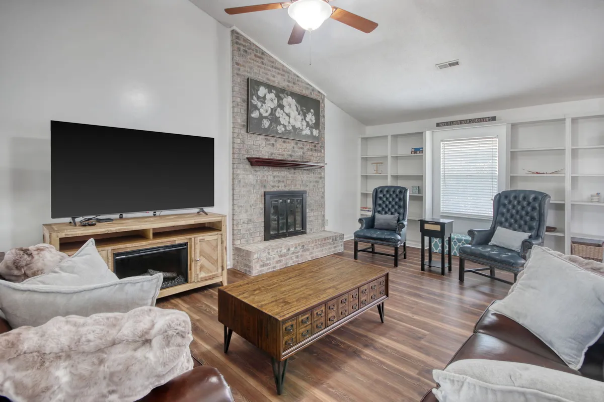 A cozy living space anchored by a brick fireplace, with a large flat-screen television, wood coffee table, and bookshelves filled with games and books. The arrangement invites group activities like movie nights and lively gatherings.