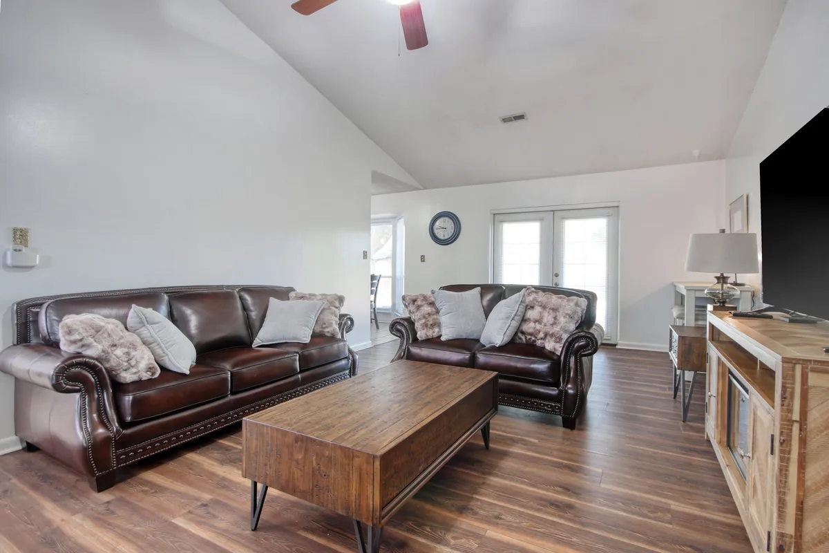 The image features a comfortable and spacious living room with leather sofas and a wood coffee table arranged for conversation. Large sliding glass doors allow ample natural light to flood the space, visually connecting the interior with the outdoors and creating an airy, welcoming atmosphere.