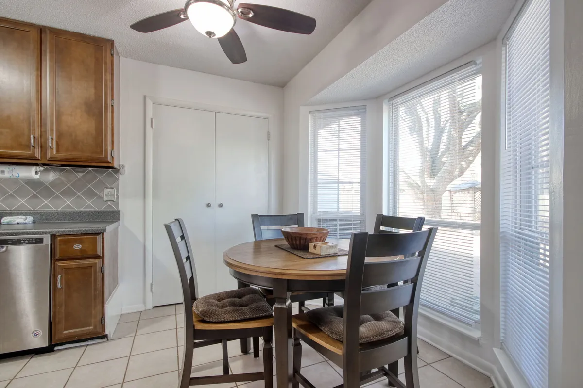 A cozy dining space situated near the kitchen, featuring a family-sized table, large windows, and kitchen cabinets in the background. The space is bathed in natural light and set up for casual or intimate meals.