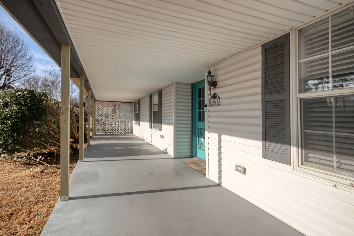 The image shows a cheerful turquoise front door with white porch railing and classic window shutters. Soft morning sunlight filters through the trees, illuminating the porch space, hinting at a welcoming entry and a spot to relax or socialize outdoors.